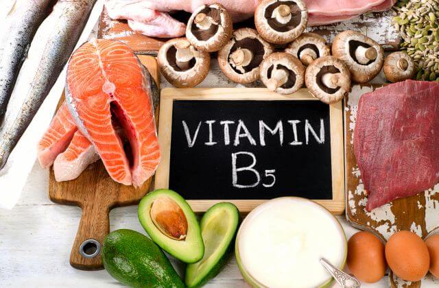 Das Bild zeigt Vitamin B5-haltige Lebensmittel wie Lachs, Avocado und Champignons. In der Mitte liegt eine Schiefertafel mit der Auffschrift "Vitamin B5".
