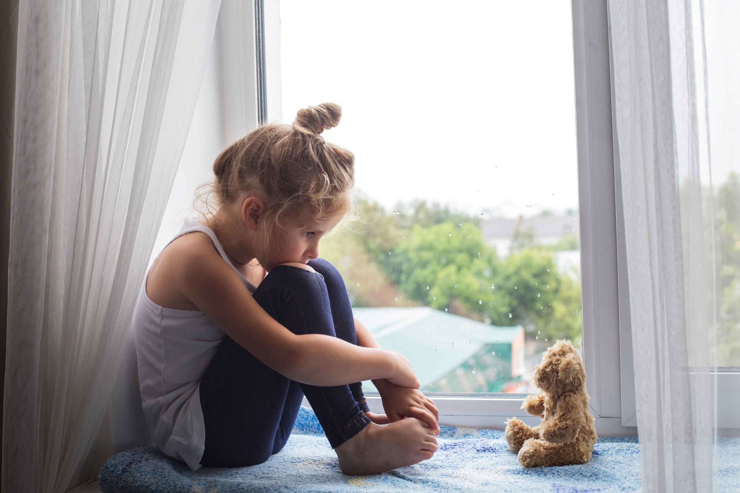 Ein kleines Mädchen sitzt niedergeschlagen am Fenster. Neben ihr sitzt ihr Teddy.