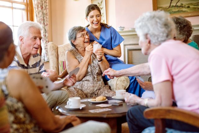 Aufnahme einer Gruppe glücklicher Senioren, die in ihrem Altersheim ein Kartenspiel spielen, während eine Krankenschwester zusieht.