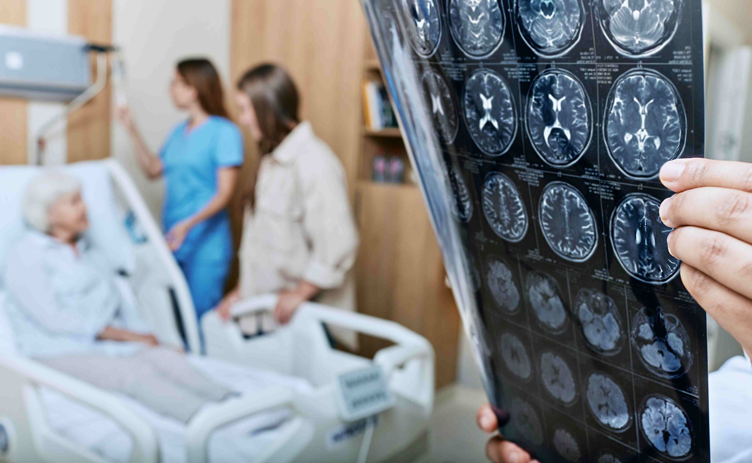 Eine alte Frau liegt im Krankenhausbett. Im Vordergrund des Bildes werden Abbildungen von Gehirnen betrachtet. Symbolbild für Schlaganfall, Demenz, und andere neurologische Erkrankungen.