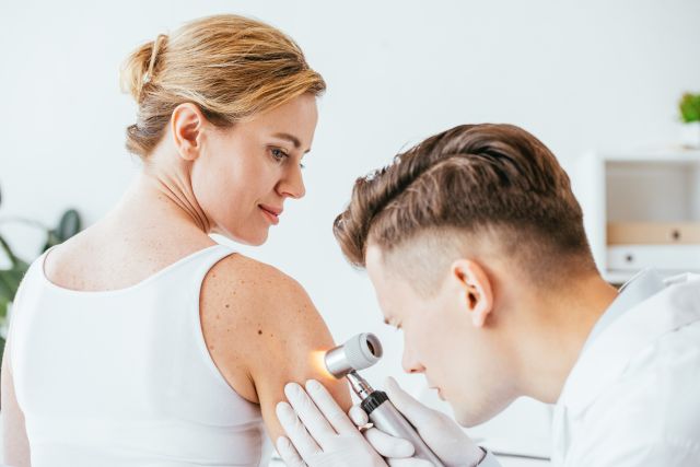 Dermatologe in Latexhandschuhen hält ein Dermatoskop in der Hand, während er eine schöne Frau mit Hautkrankheit untersucht