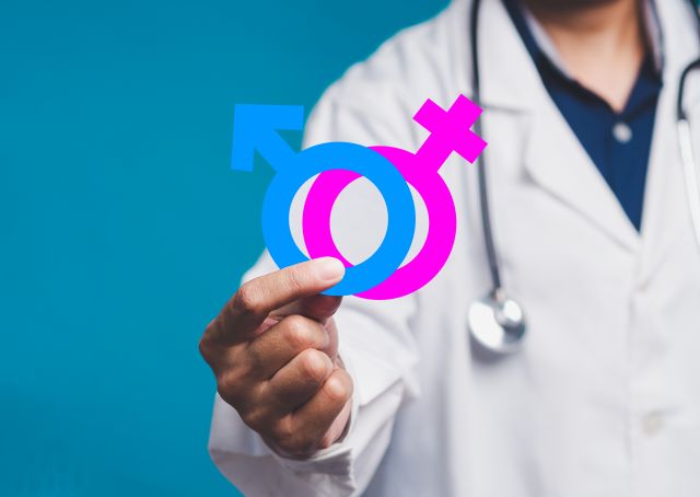 Symbolbild für Gendermedizin: Arzt hält männliches und weibliches Geschlechtssymbol vor blauem Hintergrund.