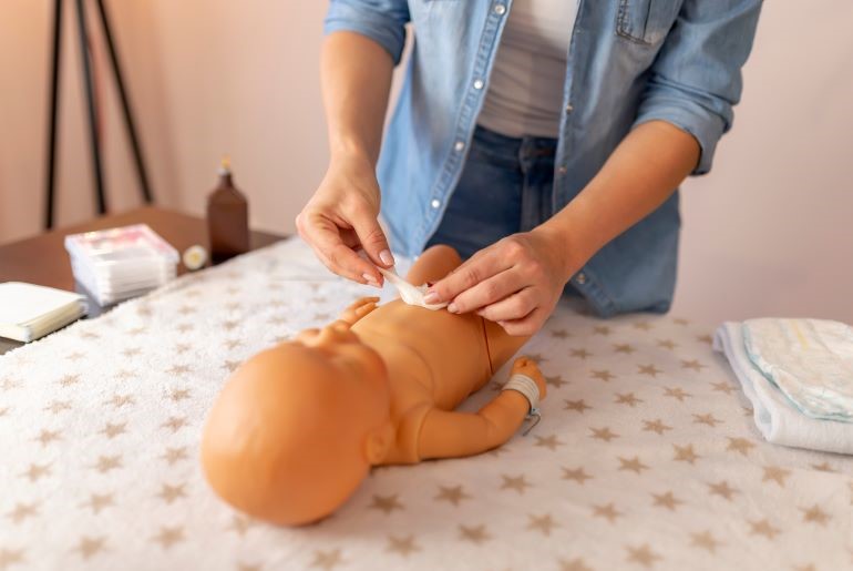 Demonstration der Nabelpflege an Neugeborenen-Puppe. Hände in Jeanshemd, Wickeltisch mit Utensilien.