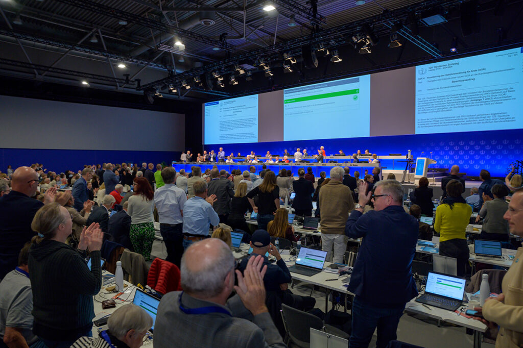 Teilnehmer einer Konferenz stehen und applaudieren nach einem Beschluss, mit einer großen Bühne und Präsentation im Hintergrund.