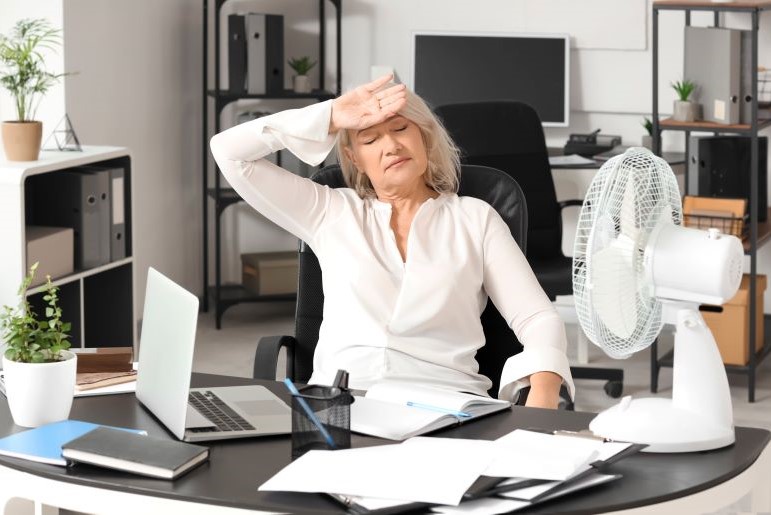 Erschöpfte Frau mittleren Alters am Schreibtisch mit Laptop und Ventilator, zeigt Symptome der Menopause bei Arbeitsbelastung.