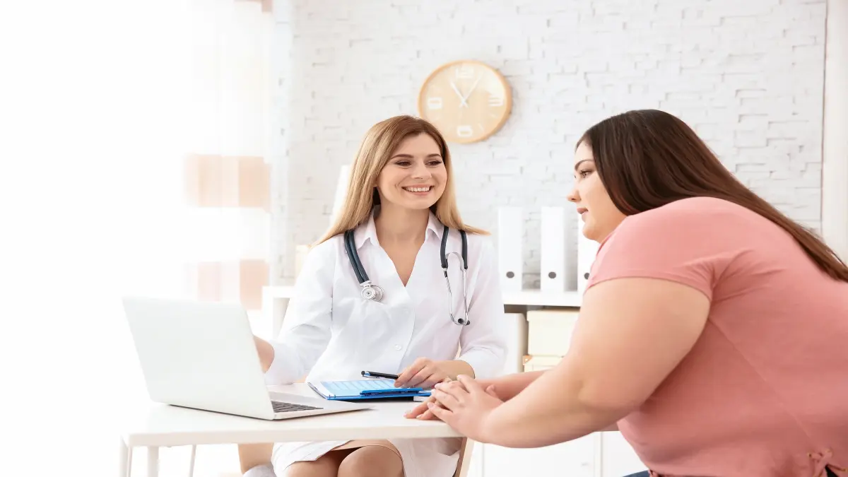 Eine Ärztin sitzt einer übergewichtigen Patientin gegenüber und zeigt auf einen Computerbildschirm, vor ihr liegt ein Klemmbrett.