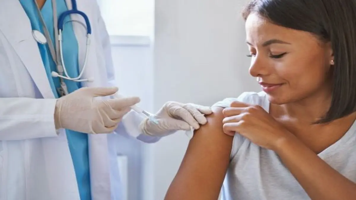 Eine Person in ärztlicher Kleidung und mit Stethoskop verabreicht einer Patientin eine Spritze in den Oberarm. Die Szene spielt sich in einem Behandlungszimmer ab. Die Patientin sitzt entspannt und hält ihren Arm locker, während das medizinische Personal mit Handschuhen die Impfung durchführt.