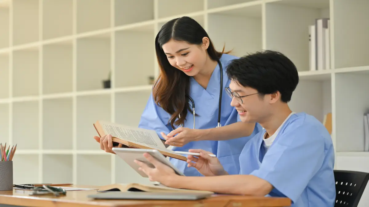 Medizinstudierende lesen in einer Bibliothek Bücher, die Studentin hält ein Buch, der Student ein Tablet.