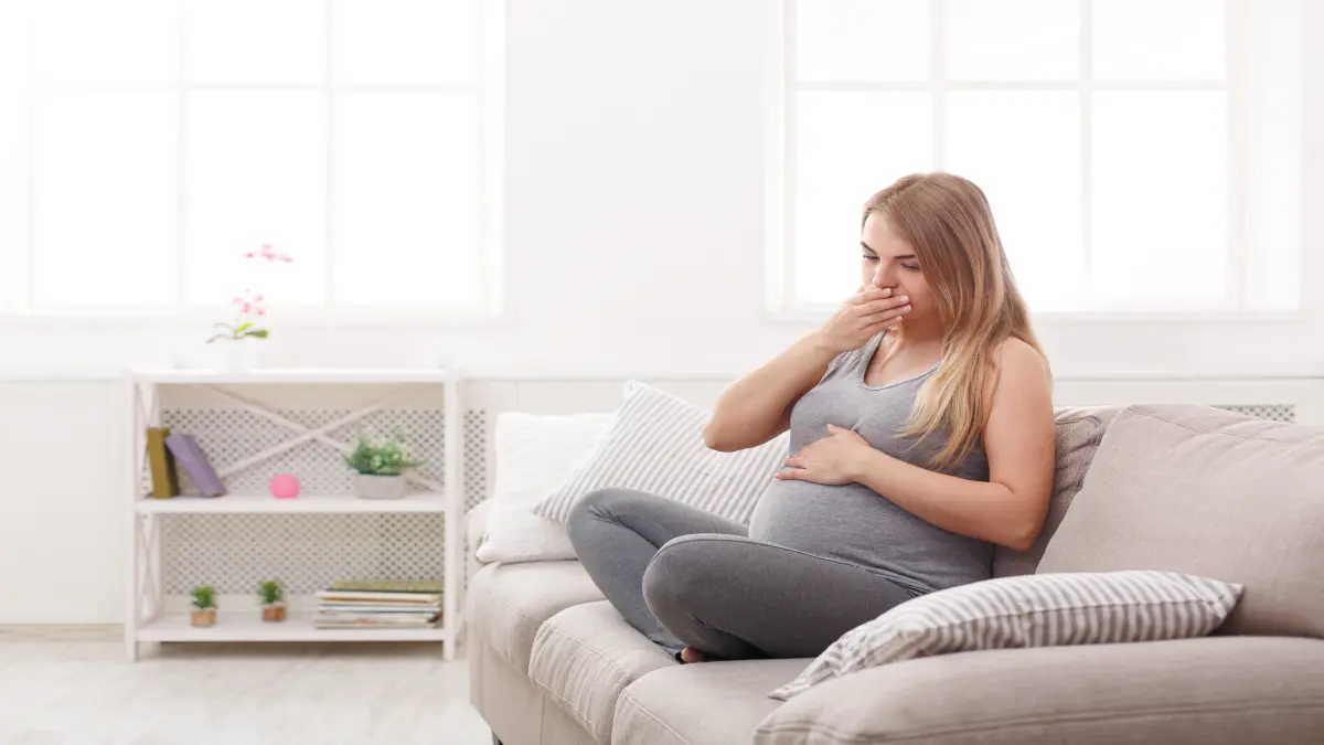 Junge Frau sitz auf dem Sofa und hält sich wegen Übelkeit die Hand vor dem Mund.