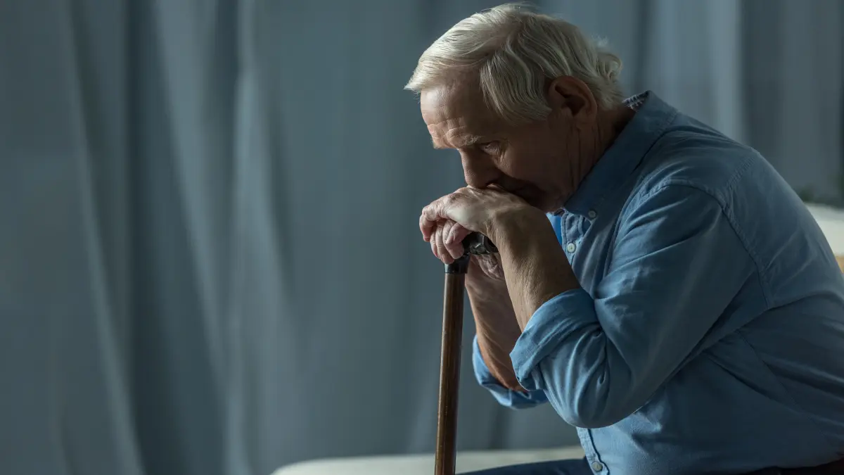 Senior sad man leans on a cane while sitting on sofa