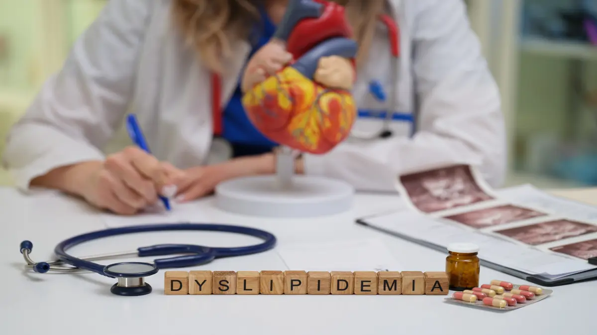 Schreibtisch mit Stethoskop, Medikamenten, Modell eines menschlichen Herzens und Holzbuchstaben, die „Dyslipidemia“ buchstabieren; im Hintergrund eine Person im Arztkittel beim Schreiben.