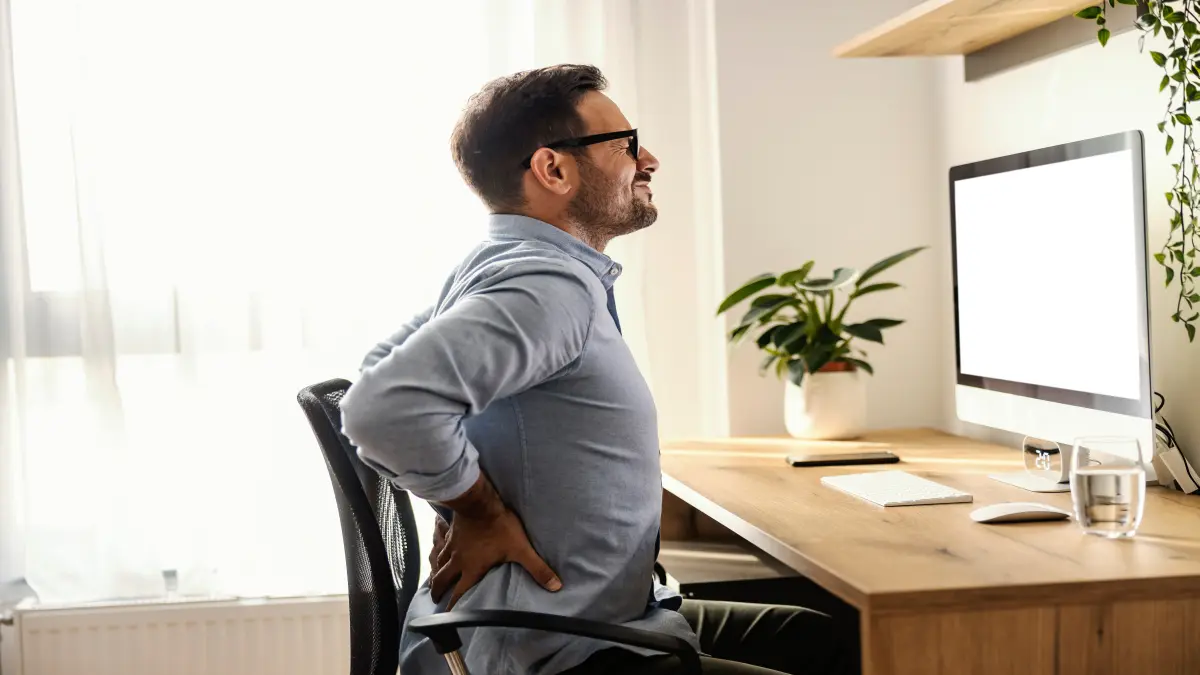 Mann sitzt am Schreibtisch und hält sich den schmerzenden unteren Rücken.