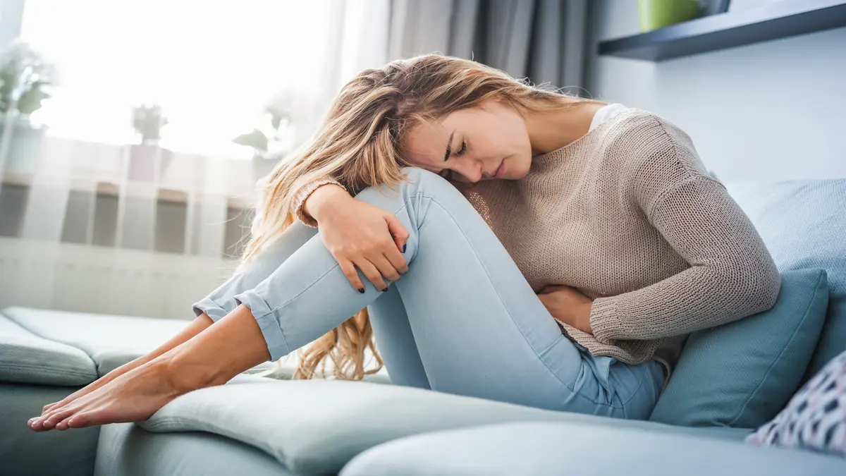 Junge Frau sitzt zusammengerollt auf dem Sofa und hält sich mit beiden Armen den Bauch, sichtbar von Bauchschmerzen oder Krämpfen geplagt.