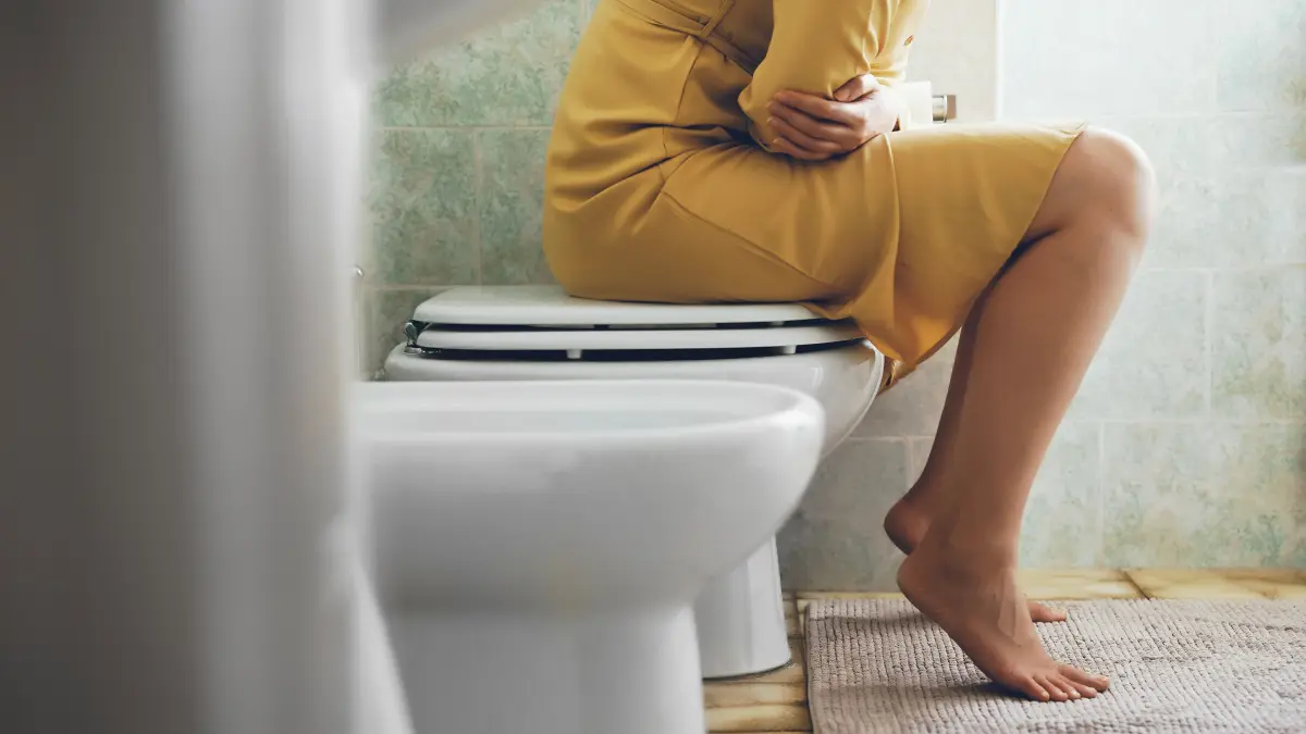 Frau sitzt mit verschränkten Armen und schmerzgeplagtem Gesichtsausdruck auf der Toilette – Symbolbild für Harnwegsinfekt und Beschwerden beim Wasserlassen.
