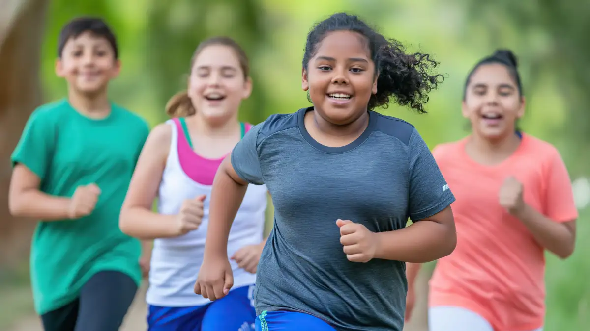 Mehrere Kinder mit Übergewicht rennen im Sportoutfit einen Straße entlang und lachen in die Kamera.