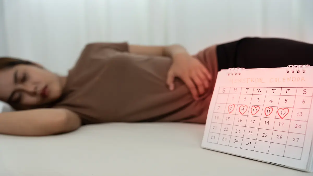 Symbolbild, Frau im Bett, die sich den Bauch hält, mit Periodenkalender im Vordergrund