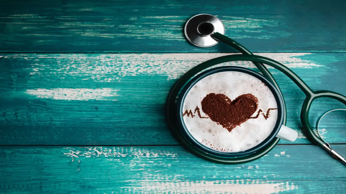 Kaffeetasse mit Herz und EKG-Linie im Milchschaum, daneben ein Stethoskop auf türkisfarbenem Holz