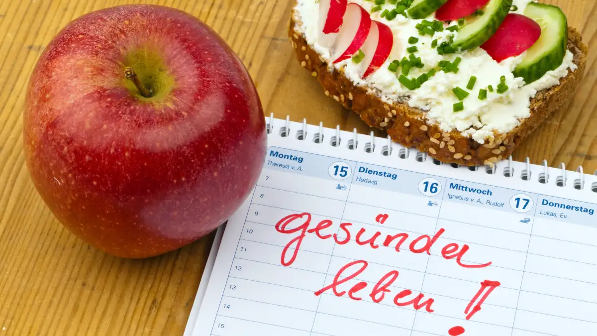 Ein Kalender mit dem roten Schriftzug "gesünder leben", daneben liegt ein Apfel sowie ein belegtes Brot mit Frischkäse und Gurke und Radieschen.