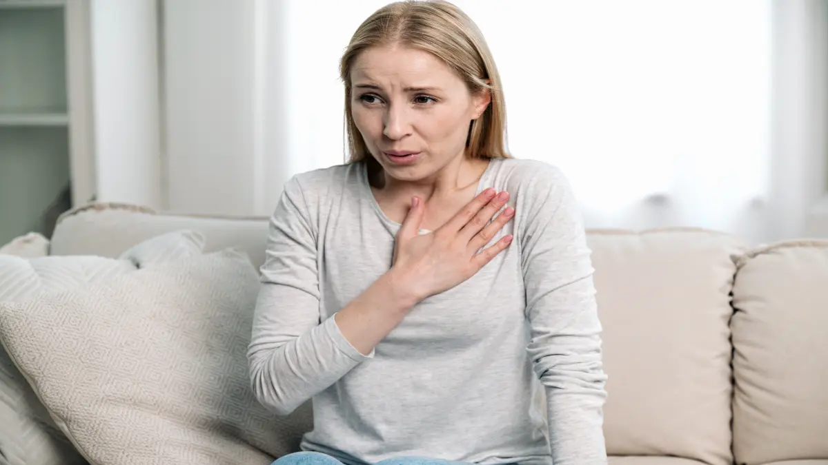 Junge Frau sitzt auf Sofa und hält sich die Brust, wirkt besorgt – Symbolbild für Atemnot.
