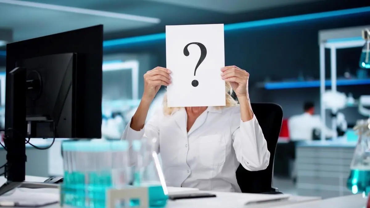 Wissenschaftlerin in einem modernen Labor hält ein Blatt Papier mit einem Fragezeichen, symbolisiert wissenschaftliche Fragen und Forschung.