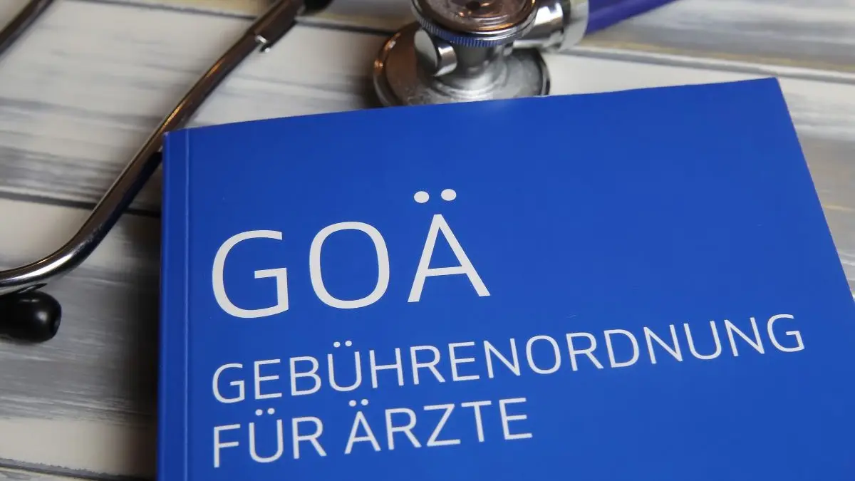 Blaues Buch mit der Aufschrift „GOÄ Gebührenordnung für Ärzte“ neben einem Stethoskop auf einem Holztisch.