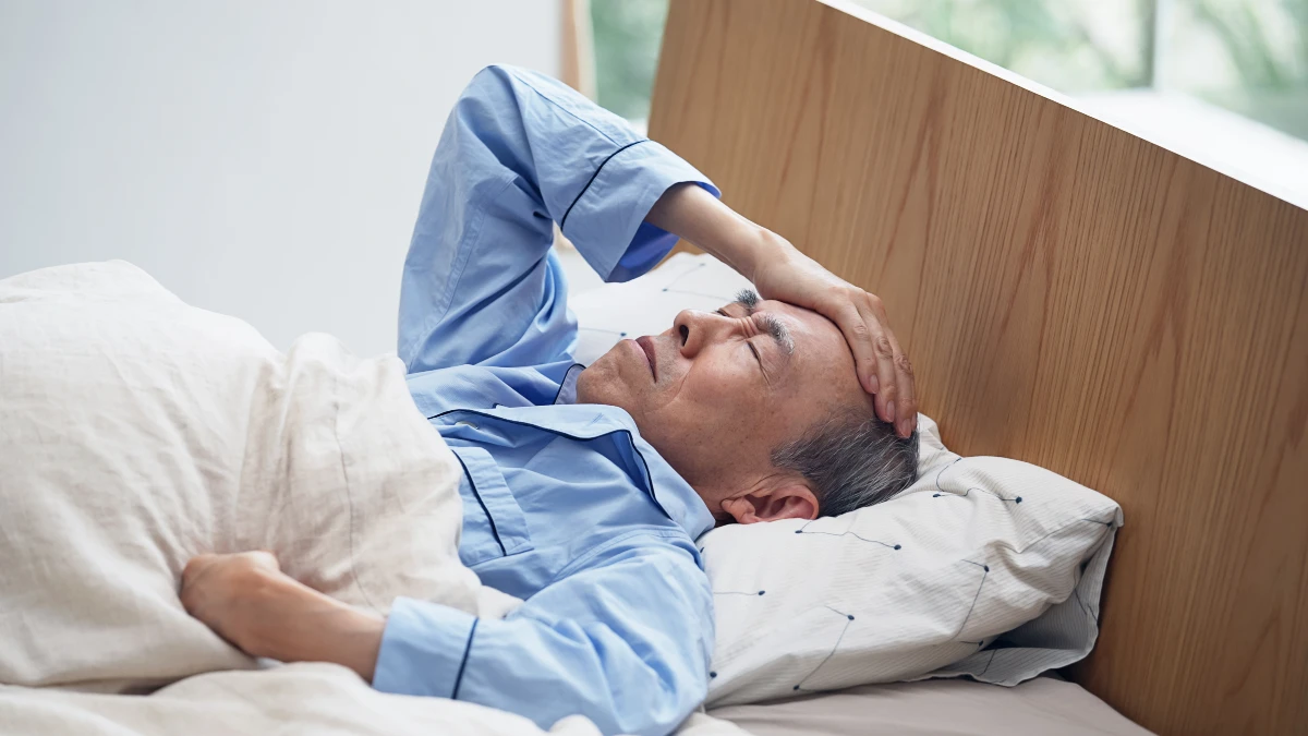 Ein alter Mann asiatischen Aussehens liegt schlaflos im Bett und hält sich den Kopf.