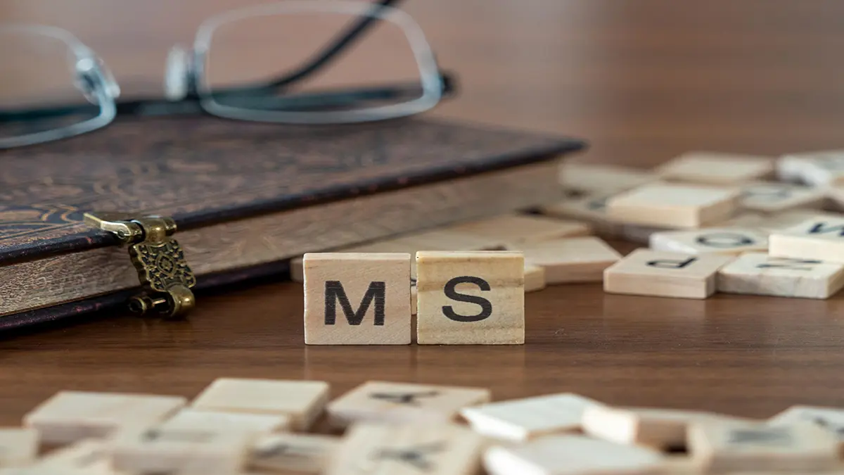 Holzbuchstaben mit der Aufschrift "MS", symbolisch für Multiple Sklerose, auf einem Tisch mit einem Buch und einer Brille.