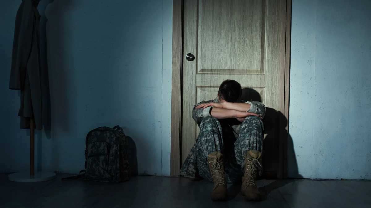 Ein Soldat sitzt in einem dunklen Zimmer, den Kopf zwischen seinen Armen verborgen. Die Atmosphäre ist bedrückend und stellt das beklemmende Gefühl einer PTBS dar.