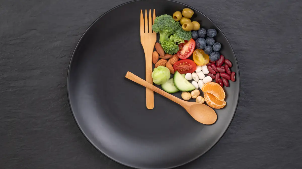 Teller mit Holzbesteck, die wie Zeiger einer Uhr auf 16 Uhr gelegt sind. Zwischen dem Besteck befindet sich eine Auswahl an Obst und Gemüse.