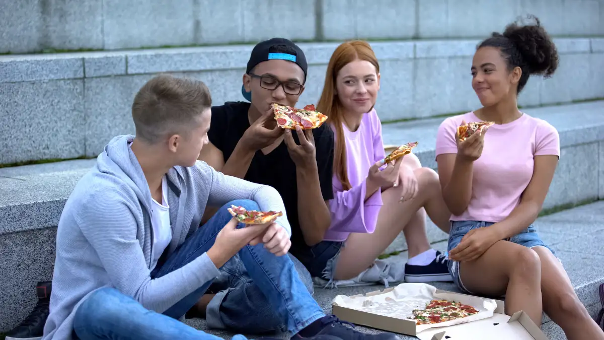 Gruppe von Jugendlichen sitzt im Freien auf Stufen und essen Pizza aus einem Pizzakarton.