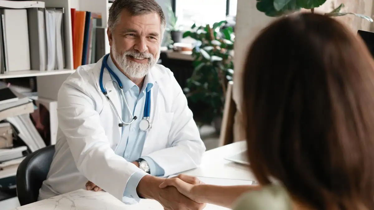 Ein männlicher Arzt sitzt an einem Schreibtisch und reicht einer gegenübersitzenden Patientin die Hand.