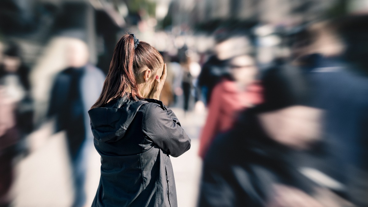 Frau mit Pferdeschwanz hält sich in einer belebten Menschenmenge die Ohren zu – Symbolbild für Angststörung, Panikattacke und Reizüberflutung