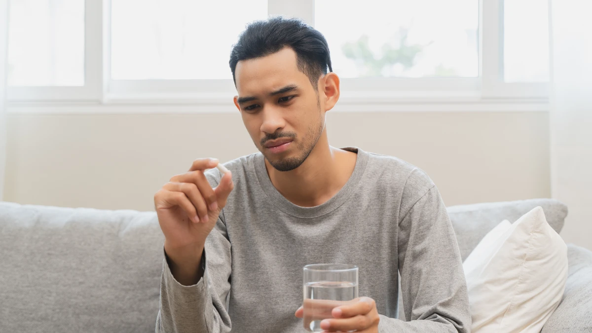 Ein Mann hält ein Glas Wasser und eine Tablette in der Hand. Er betrachtet die Tablette mit ablehnendem Blick.