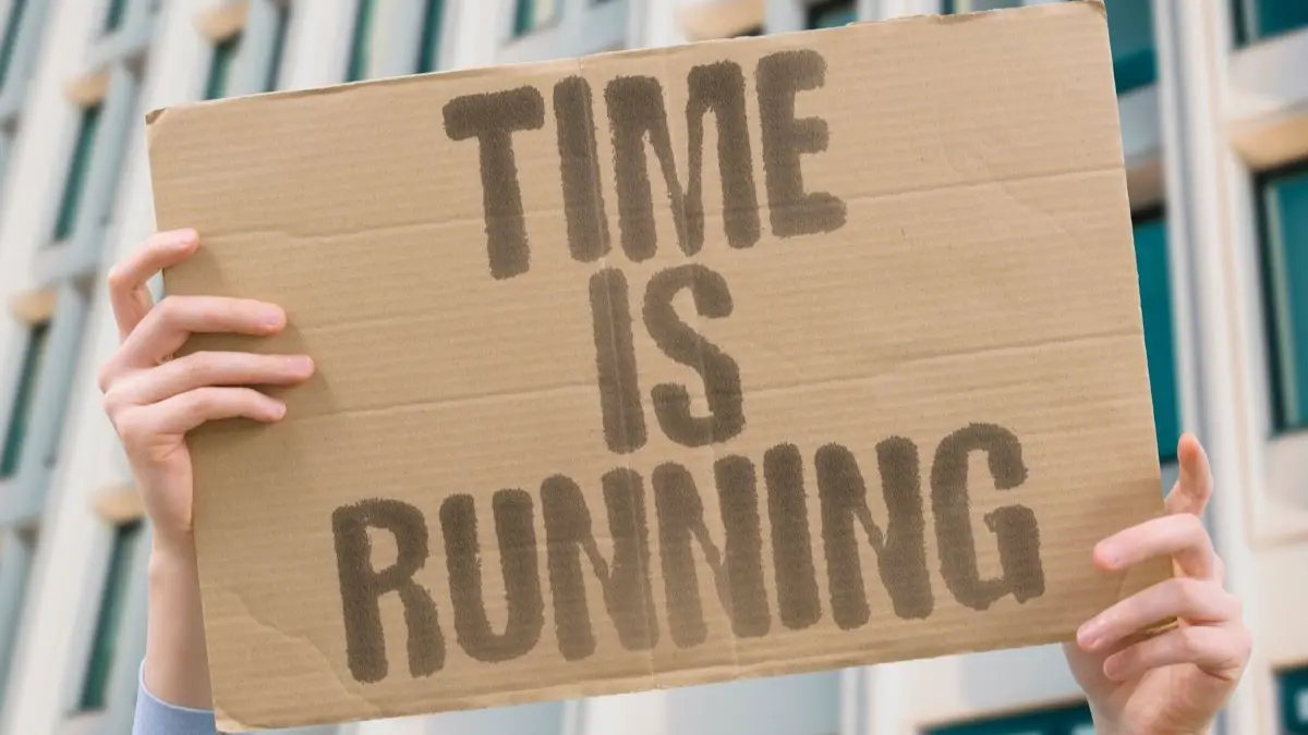 Pappschild mit der Aufschrift „TIME IS RUNNING“ wird von zwei Händen vor einem Gebäude gehalten – Symbol für Zeitdruck oder Protest