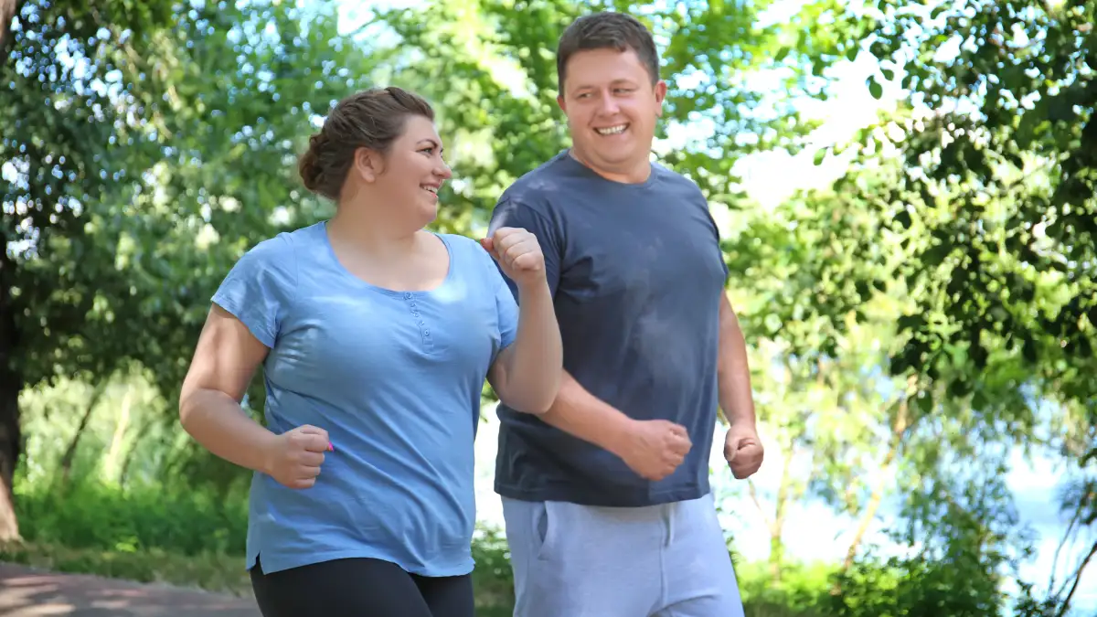 Eine Frau und ein Mann mit leichtem Übergewicht joggen in Laufkleidung in der Natur.