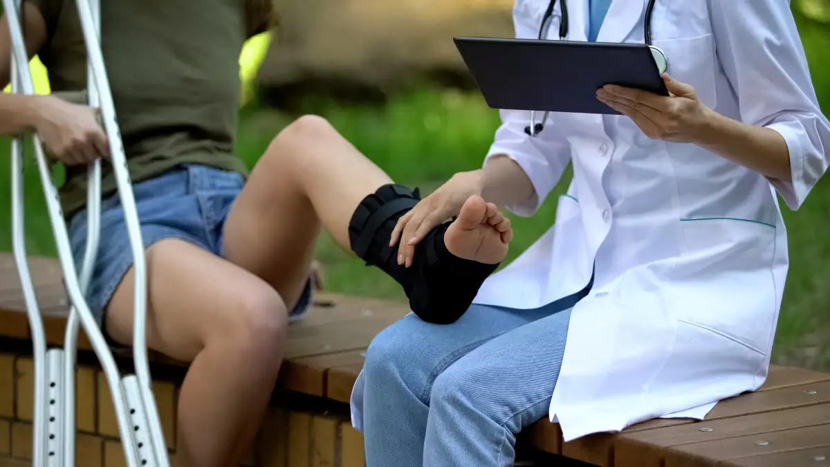Patientin mit Krücken und bandagierten Fuß sitzt auf einer Bank und Ärztin mit Tablet sitzt daneben.