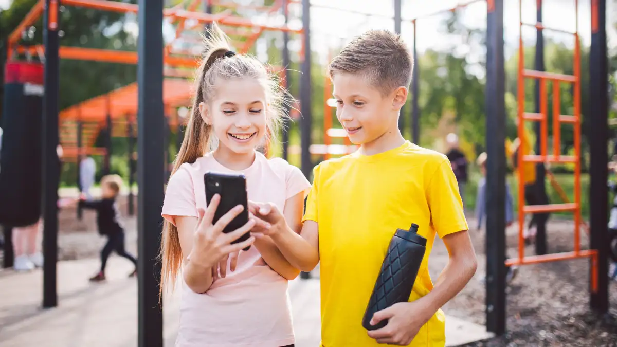 Ein Mädchen und ein Junge im Grundschulalter stehen lächelnd auf einem Spielplatz und schauen gemeinsam auf ein Smartphone.