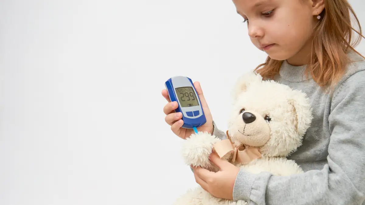 Mädchen misst bei einem weißen Plüschteddy im Arm im Spiel den Blutzuckerspiegel mit einem Messegrät.