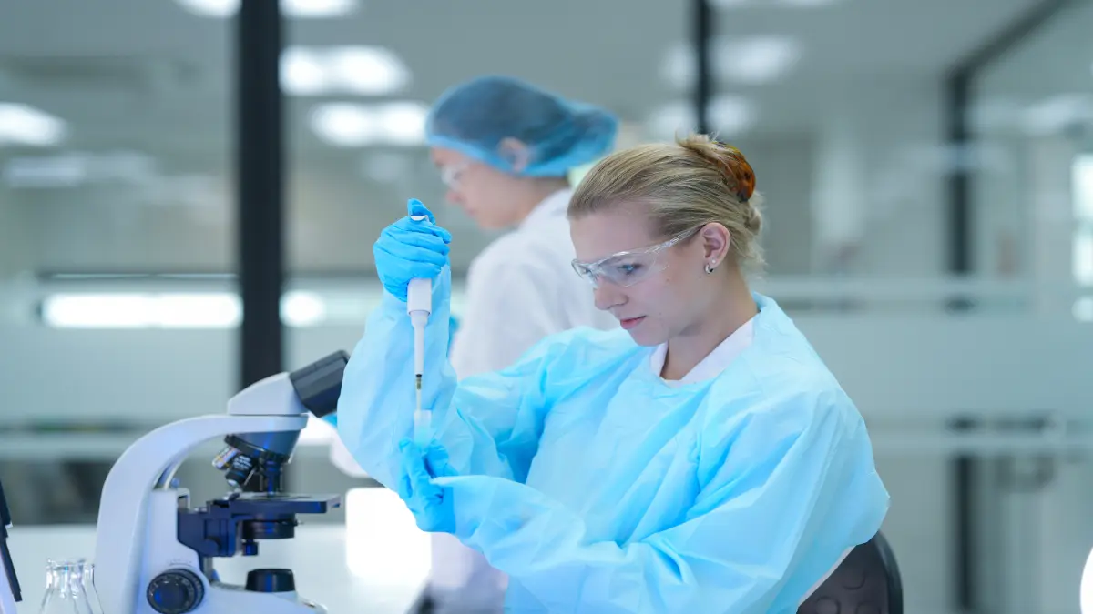 Wissenschaftlerin in Schutzkleidung pipettiert eine Probe im medizinischen Labor, im Hintergrund eine weitere Forscherin