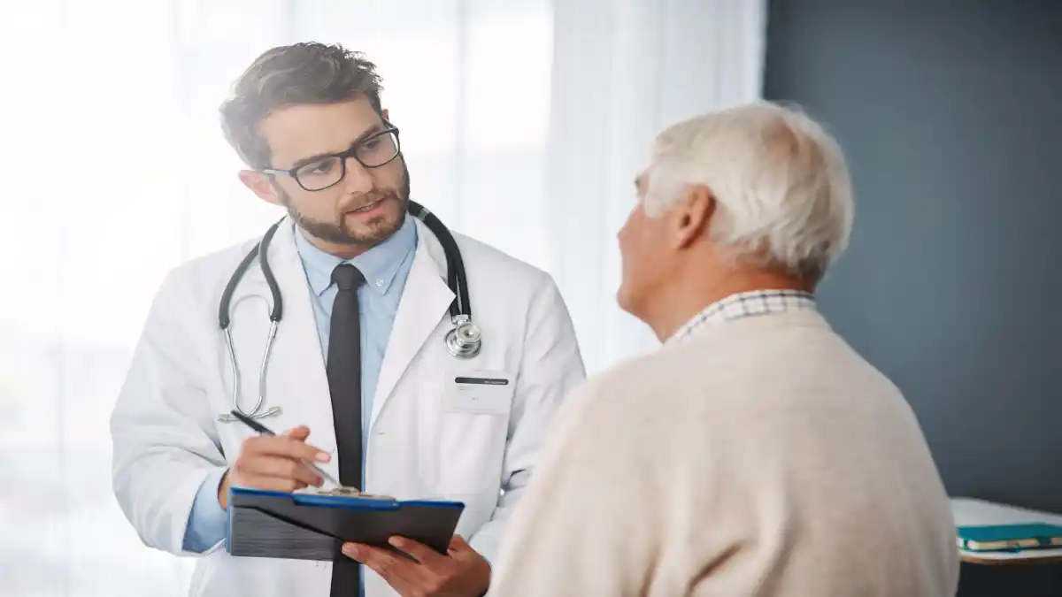 Auf dem Foto hält ein junger Arzt eine Klemmbrett und spricht mit einem älteren Patienten.