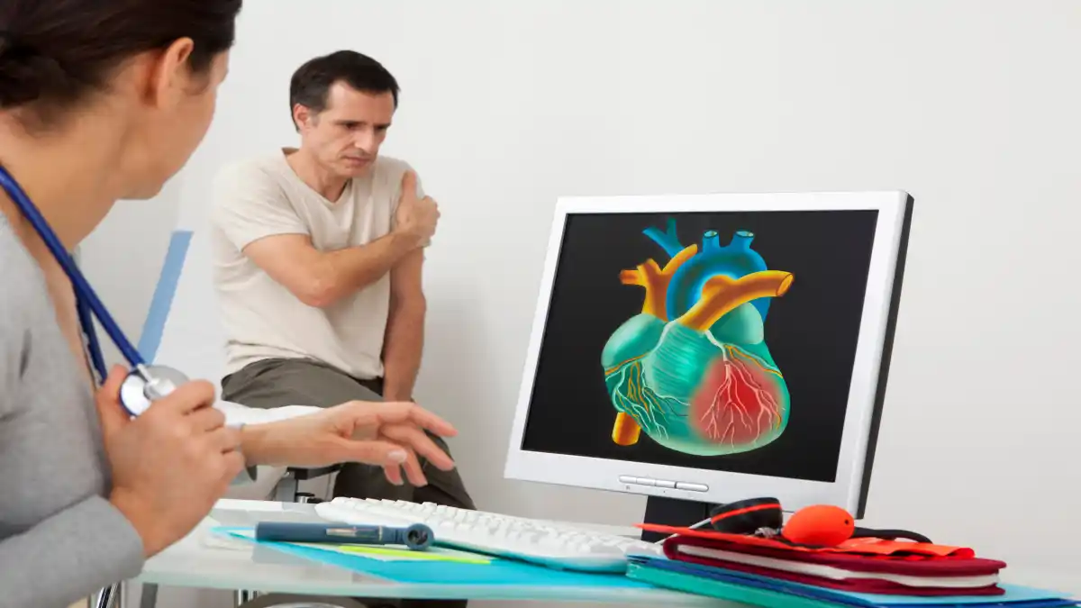 Man sieht auf dem Bild einen Mann, der sich den linken Arm hält. Im Vordergrund sitzt eine Ärztin auf einem Tisch und vor ihr steht ein Computer auf dessen Bildschirmmonitor ein 3D-Herz zu sehen ist, in dem die Gefäße rot markiert sind.
