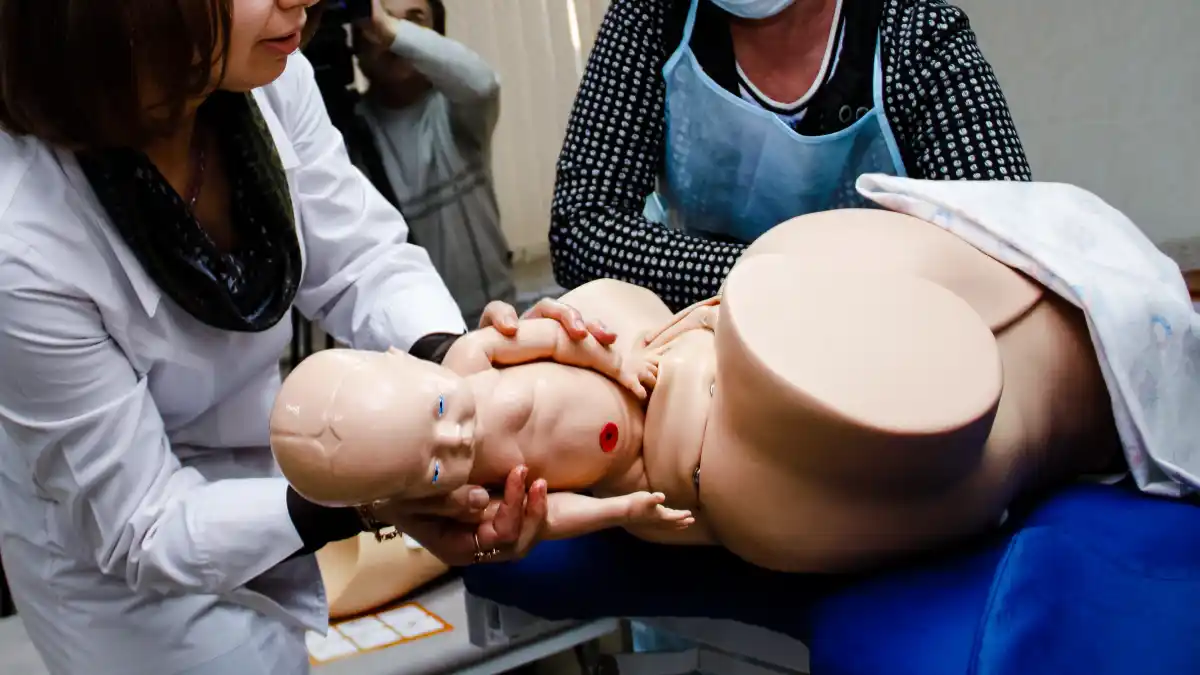 Hier wird die Simulation einer Geburt mithilfe einer Simulatorpuppe gezeigt, hinten steht ein Mann mit Kamera.