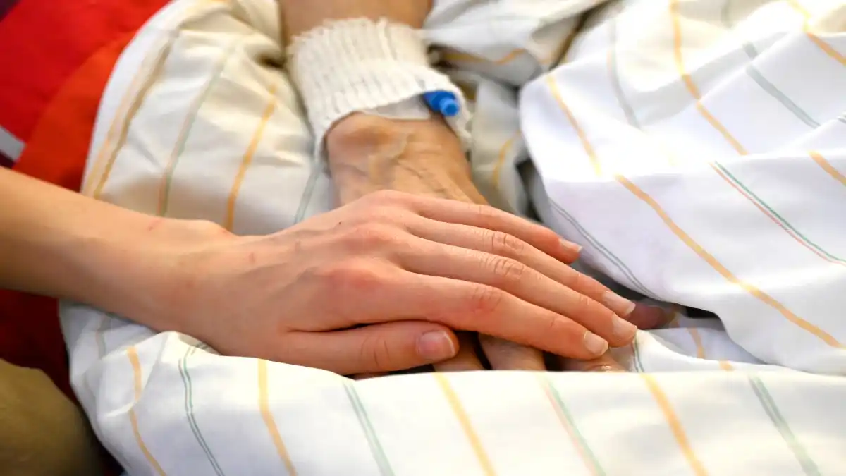 Das Bild zeigt wie jemand die Hand einer im Krankbett liegenden Person hält.