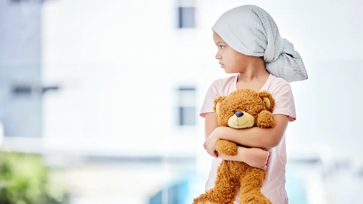 Mädchen mit Leukämie trägt ein Kopftuch und hält einen Teddybär