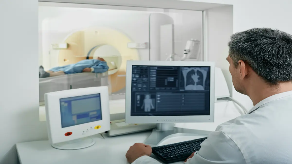 Radiologe wertet CT-Aufnahmen der Lunge am Monitor aus, im Hintergrund Patient im CT-Scanner