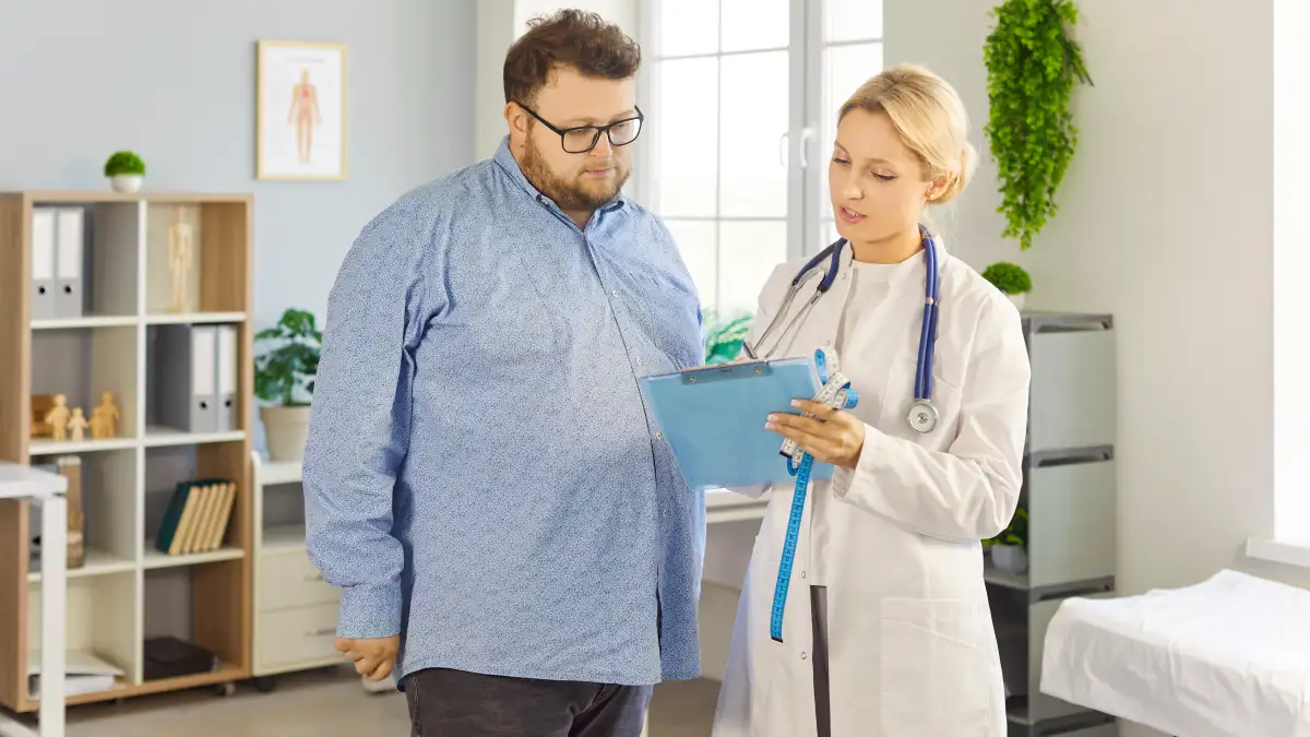Eine Ärztin steht neben einem adipösen Mann und erklärt Untersuchungsergebnisse auf einem blauen Klemmbrett.