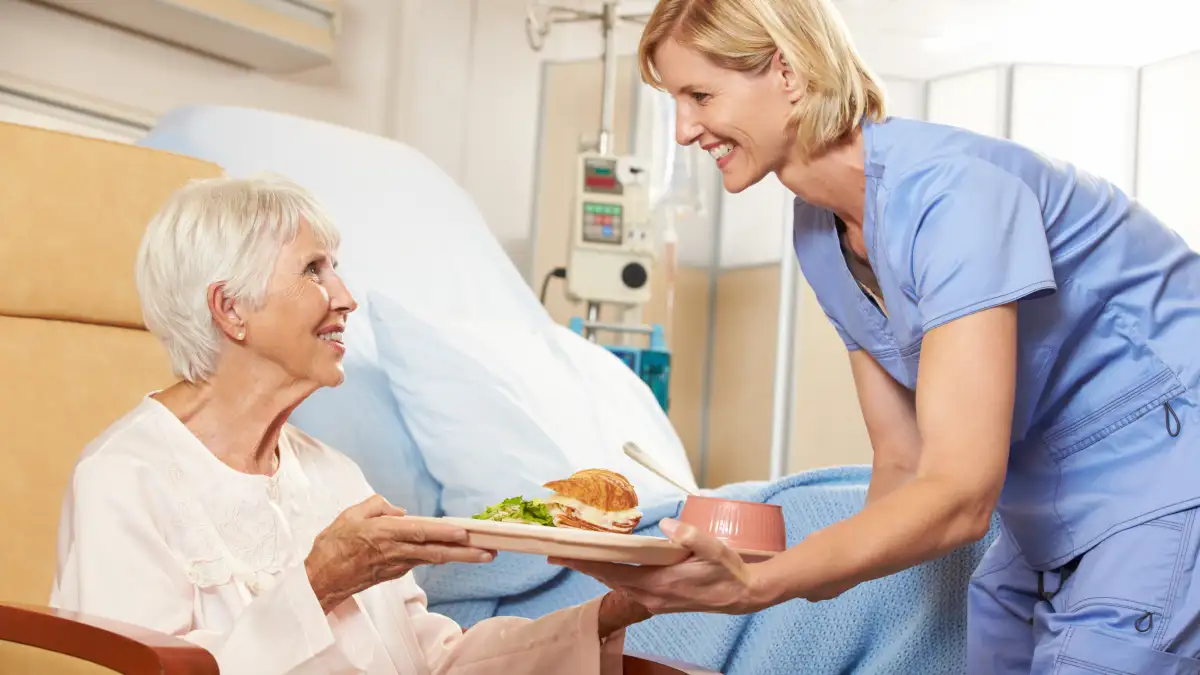 Eine ältere Dame erhält neben ihrem Krankenhausbett ein Tablett mit Essen von einer Krankenschwester im blauen Kasak.