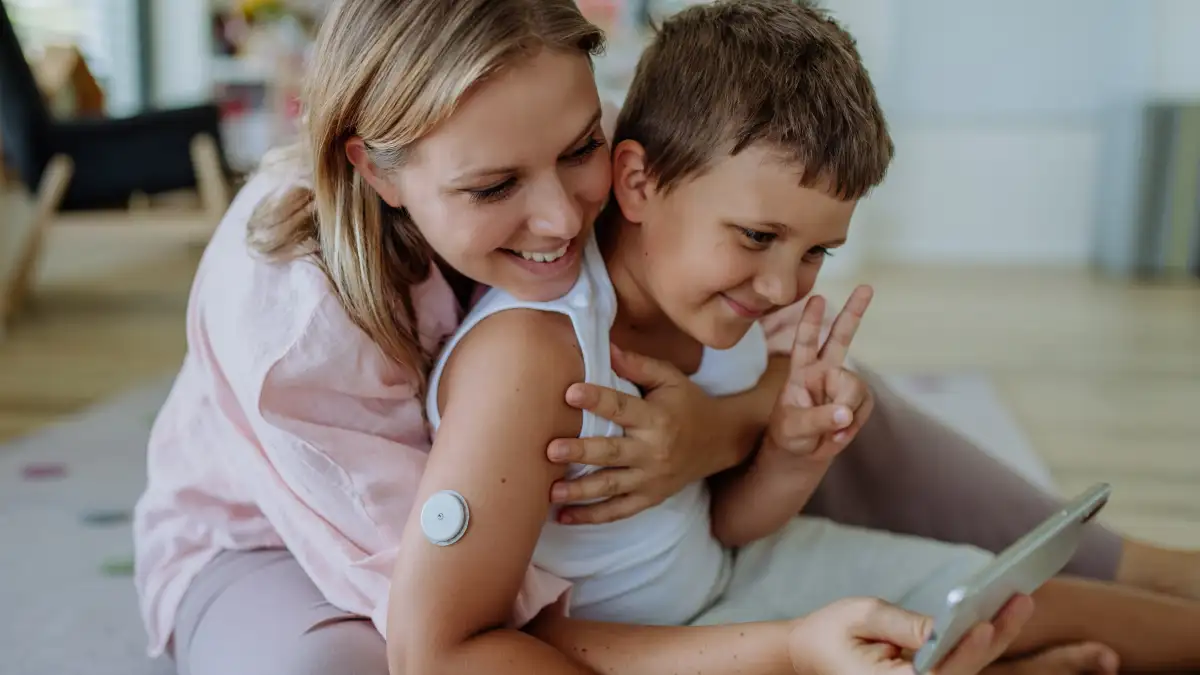 Eine Mutter umarmt ihren Sohn, der einen Glukosesensor am Arm trägt, von hinten und beide lächeln in ein Smartphone.