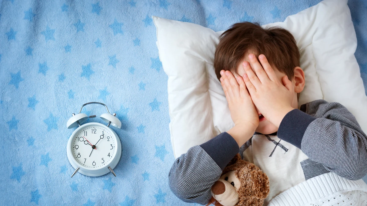 Kind liegt im Bett, hält sich die Hände vors Gesicht, neben ihm ein Wecker – Symbol für kindliche Insomnie
