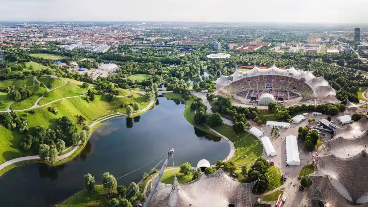 Luftaufnahme vom Olympiapark in Muenchen mit Stadion, Seen und Gruenflaechen als Hinweis auf FOBO 2026 in Muenchen